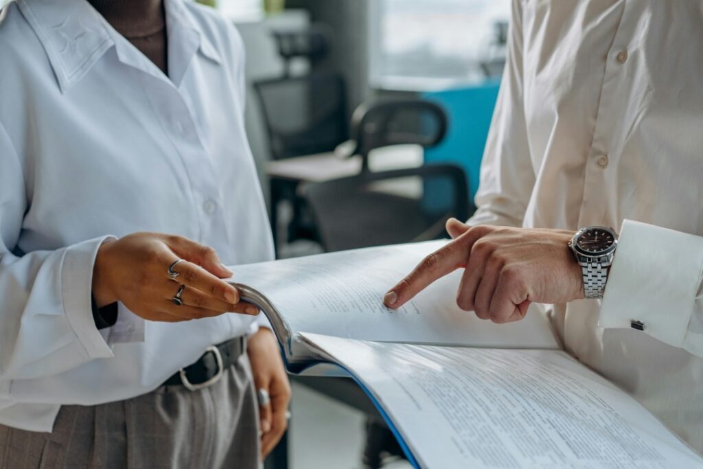 Two people dressed in business examining paperwork. One holds the top of the page as the other points at something on the page.