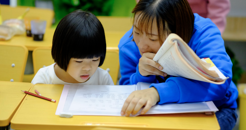 a teacher leaning over a workbook with a young child and holding a doubled-over book