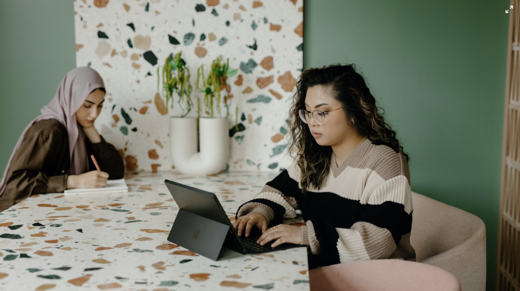 Two women sat at a decorative table, one working on a tablet, one writing in a notebook