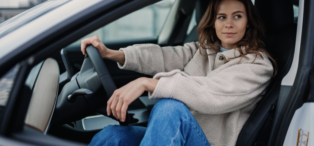 woman sat back in a car with her hands on the steering wheel, looking off into the middle distance 