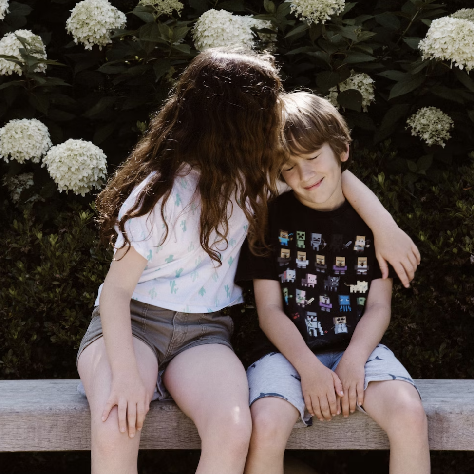 an older child with long hair embracing a younger child with shorter hair who looks upset.