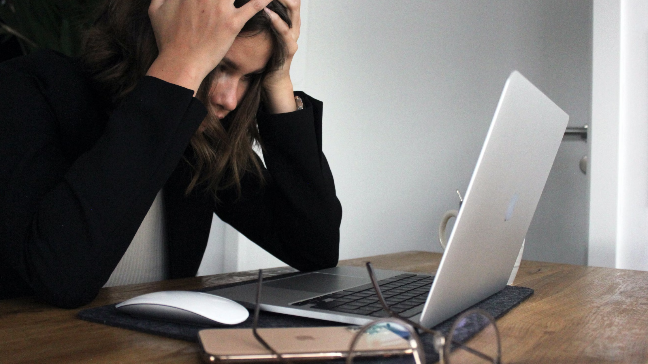 woman with short dark hair sat at a laptop, head in her hands, a pair of glasses to her side 