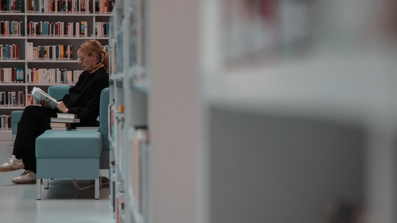 a side view of a woman sat in a library on a blue chair, legs crossed, a book open in her lap