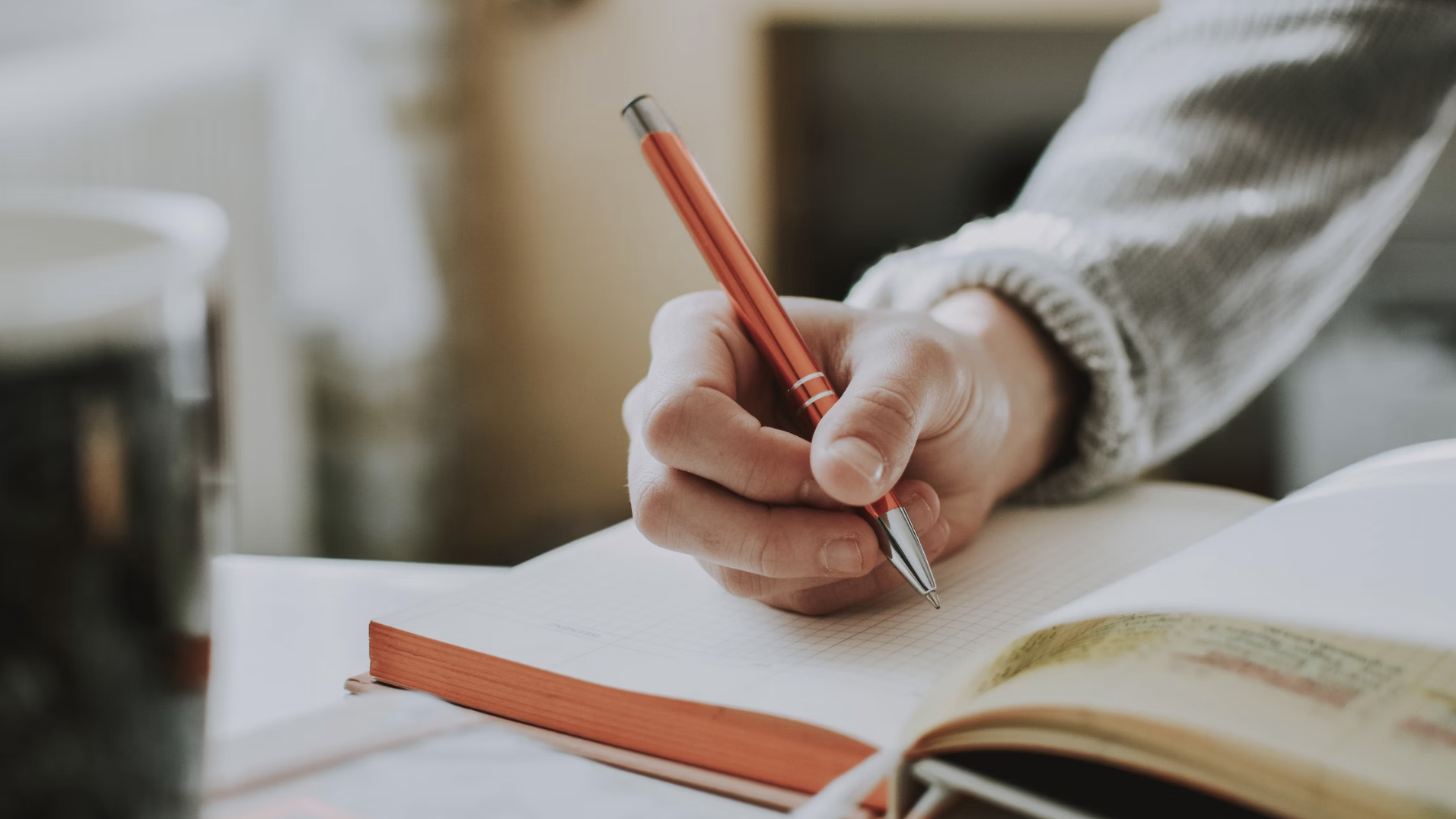 an orange pen in a hand, held above a notebook with squared pages.
