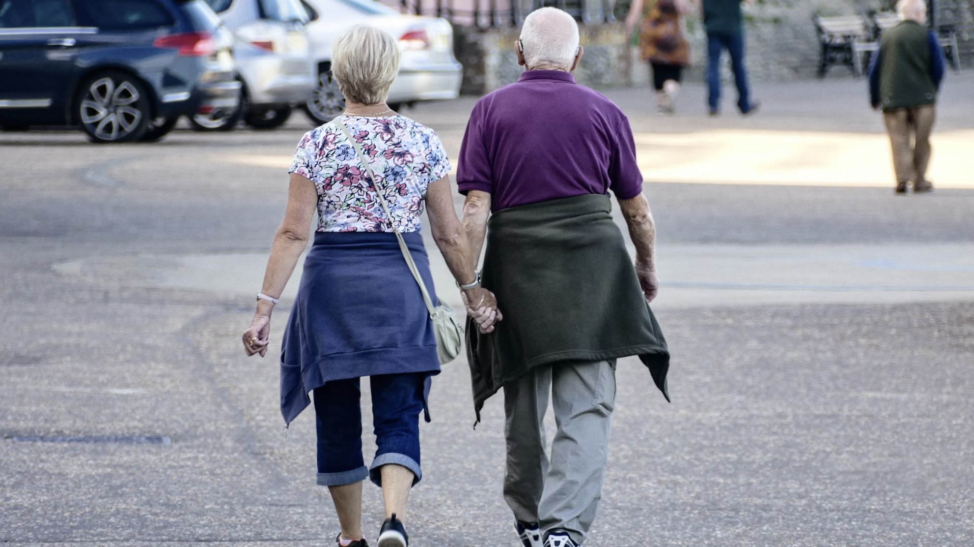 two older people walking away from the camera, dressed casually, holding hands 