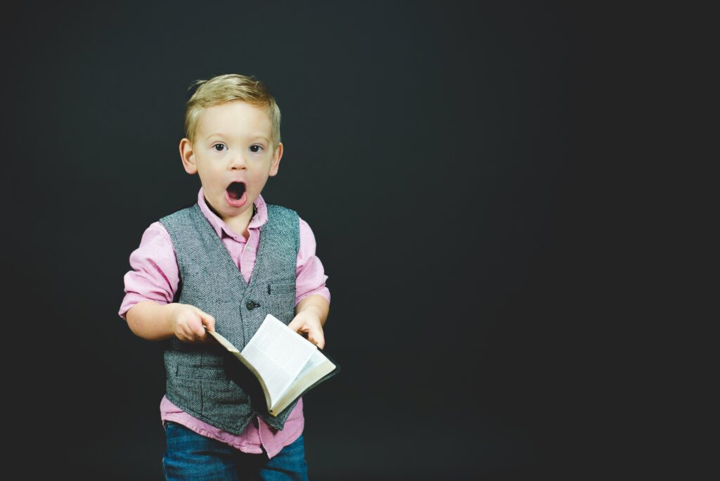 A child with short blonde hair holding an open book and looking shocked. He's wearing a smart grey waistcoat and a pink collared short. 