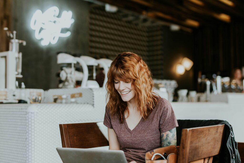 A young person sitting at a laptop in a coffee shop, smiling down at it. They have medium-length red hair and a sleeve tattoo. 