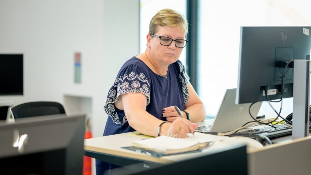 an older person in glasses and a blouse sat in an office, behind a monitor, writing in a notebook