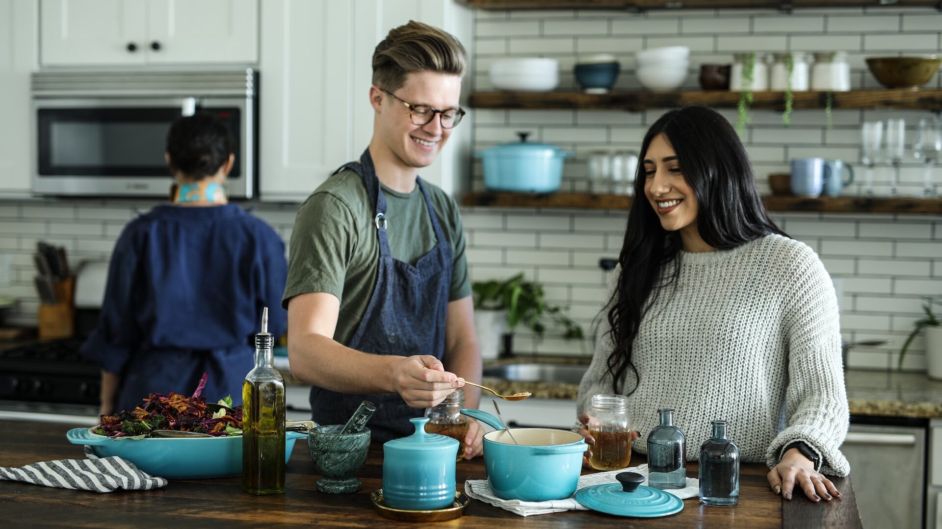 three people cooking in a modern tiled kitchen, one facing away, one adding a spoonful of ingredients to a blue pot, and one with long hair looking on with a smile.