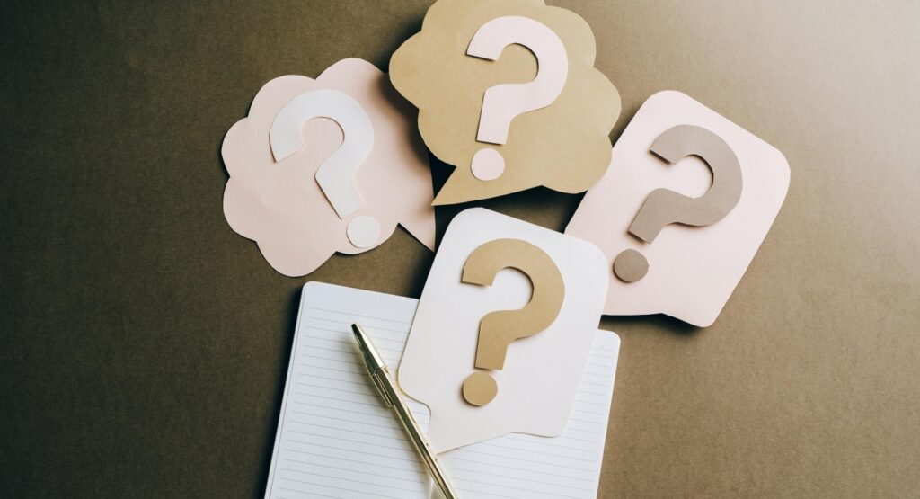 An open notebook, a gold-coloured pen and a lot of post-it notes with question marks on them, spread out on a beige desk background. 