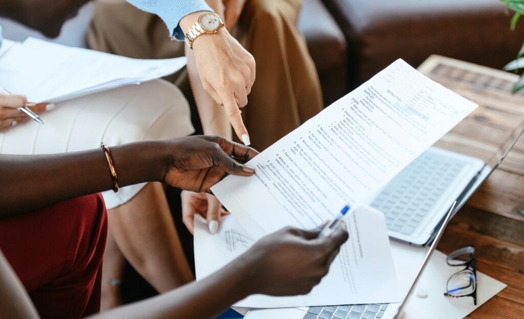 One pair of hands holding a document, another, wearing a watch, pointing at the bottom part of the document. There are open laptops on the table behind.