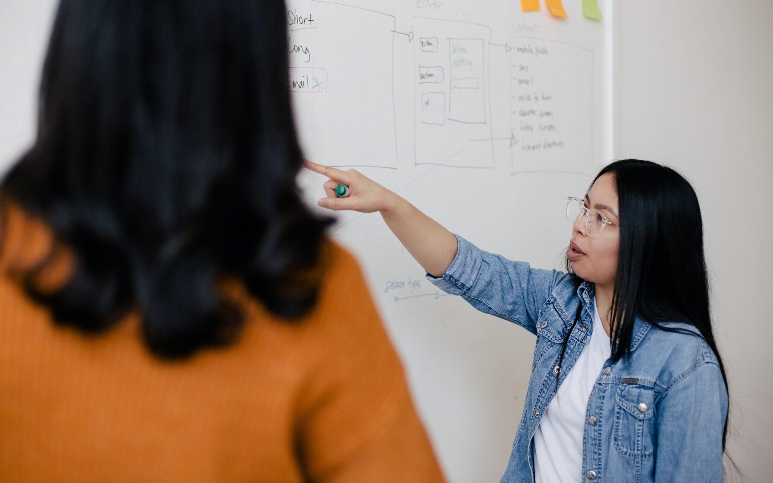 woman in glasses pointing at a whiteboard 