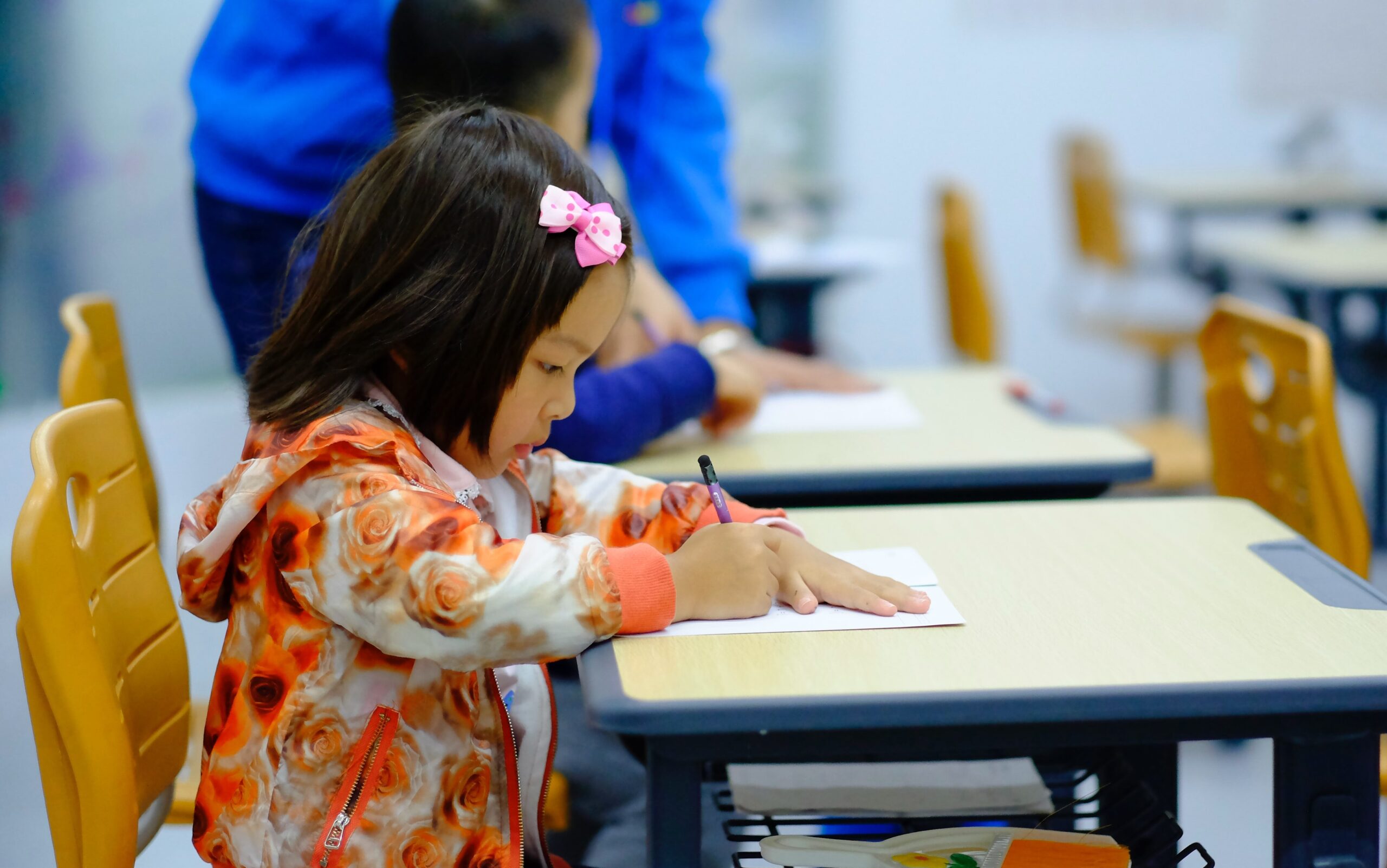 younger child with pink bow in hair, focused on written work and sat at a desk