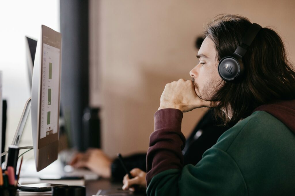 An employee with facial hair and long hair wearing heapdhones, holding their chin and looking at a screen. 