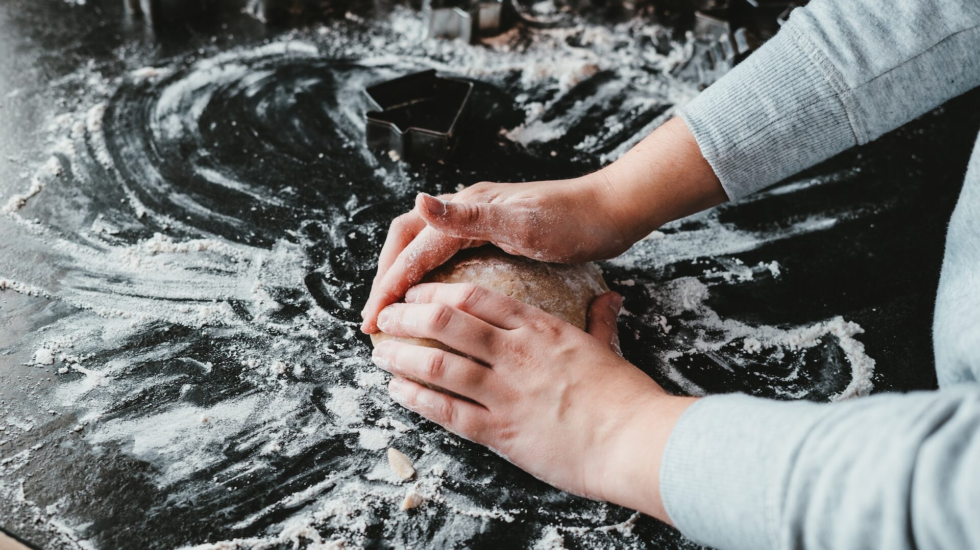 two hands holding a ball of bread dough against a floured black worksurface.