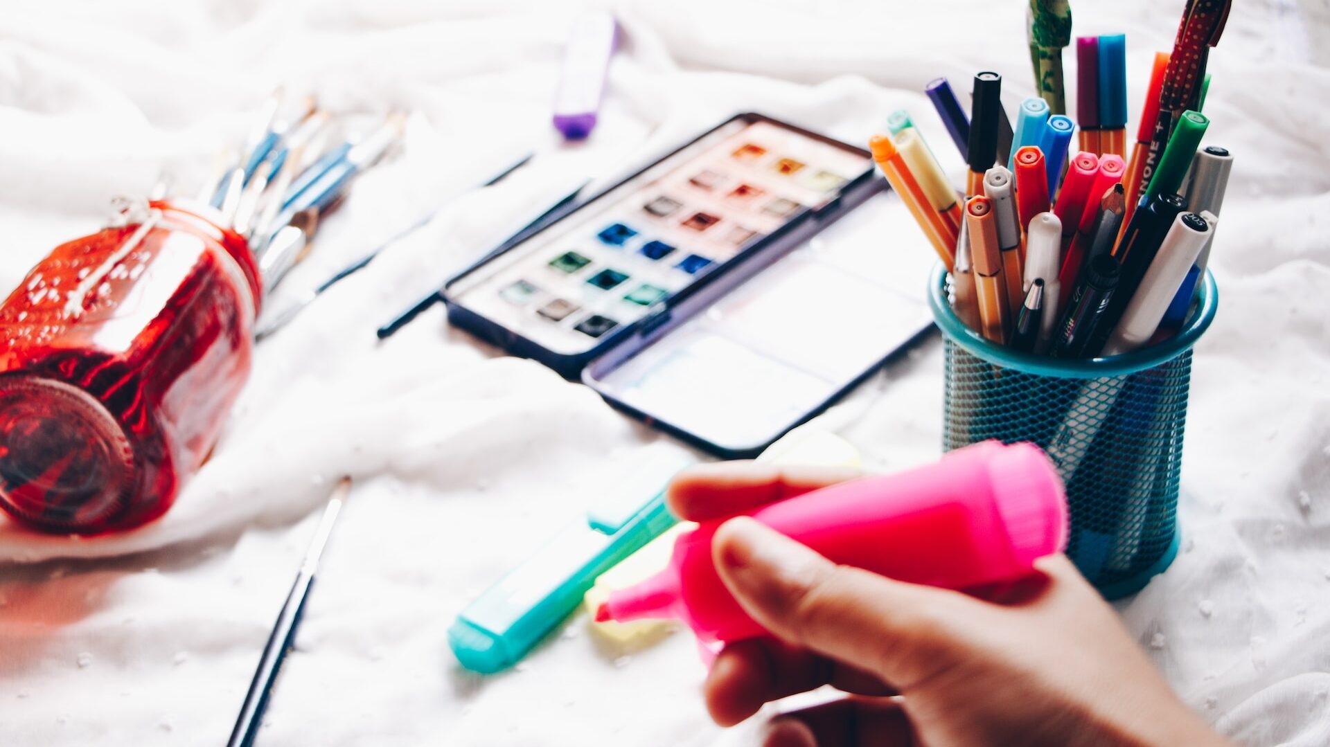 An assortment of writing implements— an out of focus hand in the foreground holds a pink highlighter, with a wire pot and a tipped glass pot of assorted pens and pencils behind. There is also a paint set. 