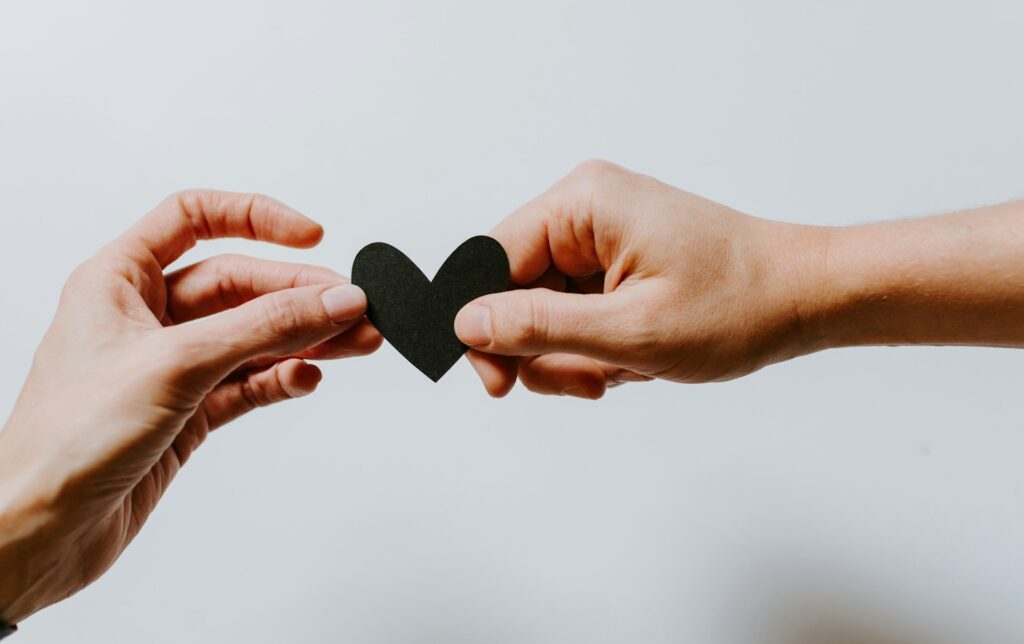 Two hands reaching out, both holding a black cut-out paper heart. 