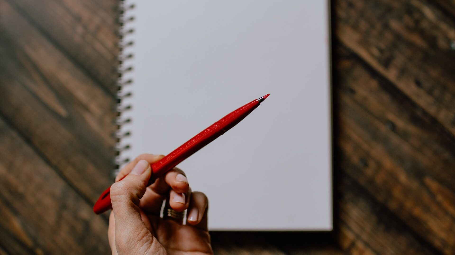 A manicured hand holding a red felt tip pen above a blank white page in a spiral bound notebook, on a dark wooden countertop. 