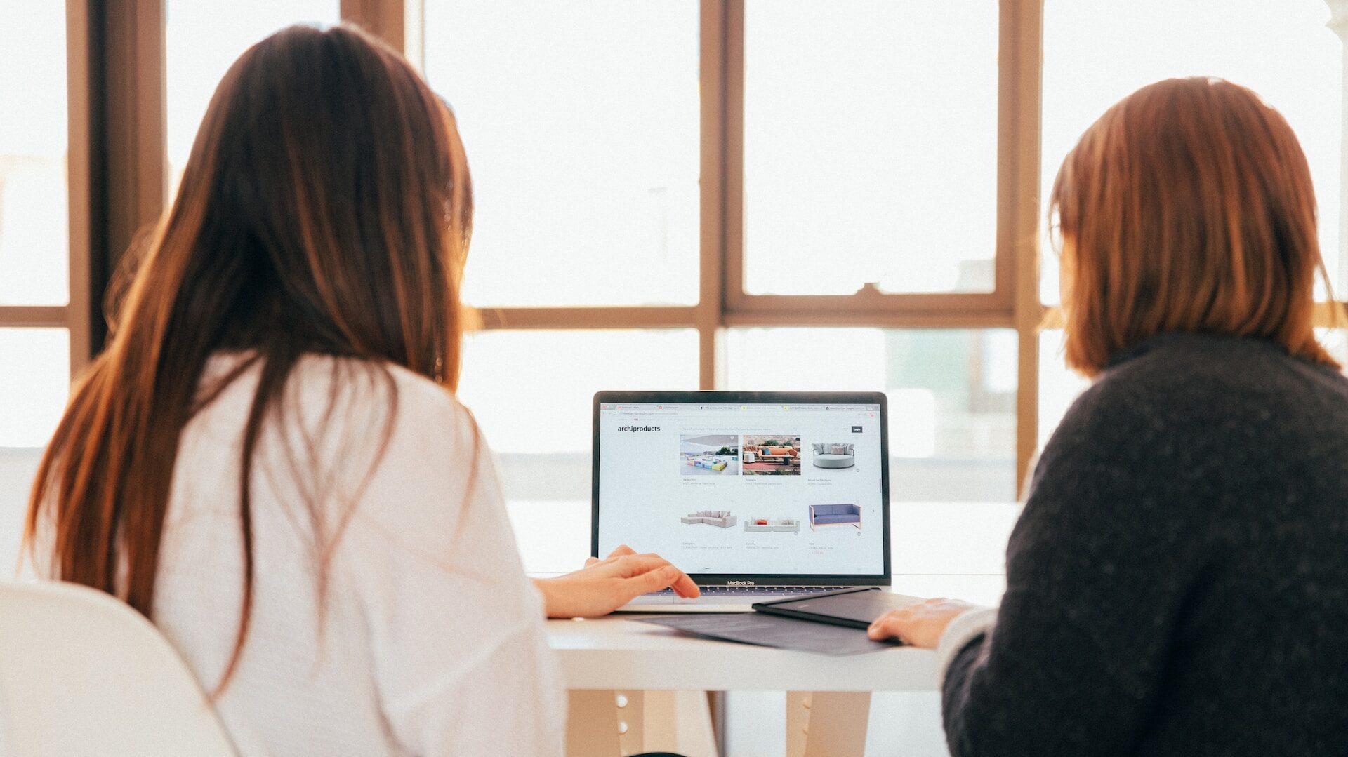 Two women collaborating on a project in front of an open laptop. Both are brunette, one with a bob, one with back-length hair. 