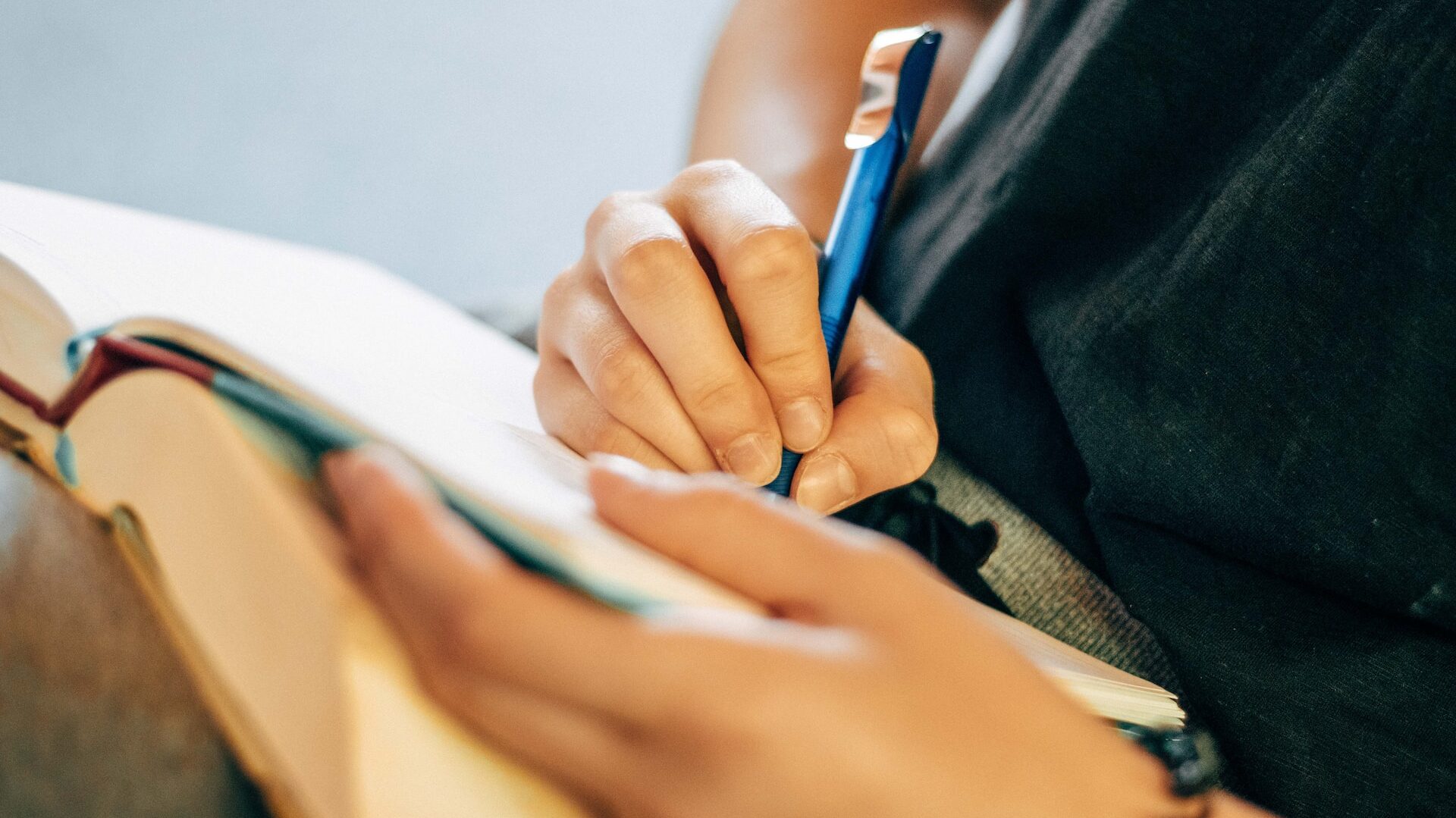 a pair of hands writing in a notebook, holding a light blue pen as the notebook is propped on their knees. 