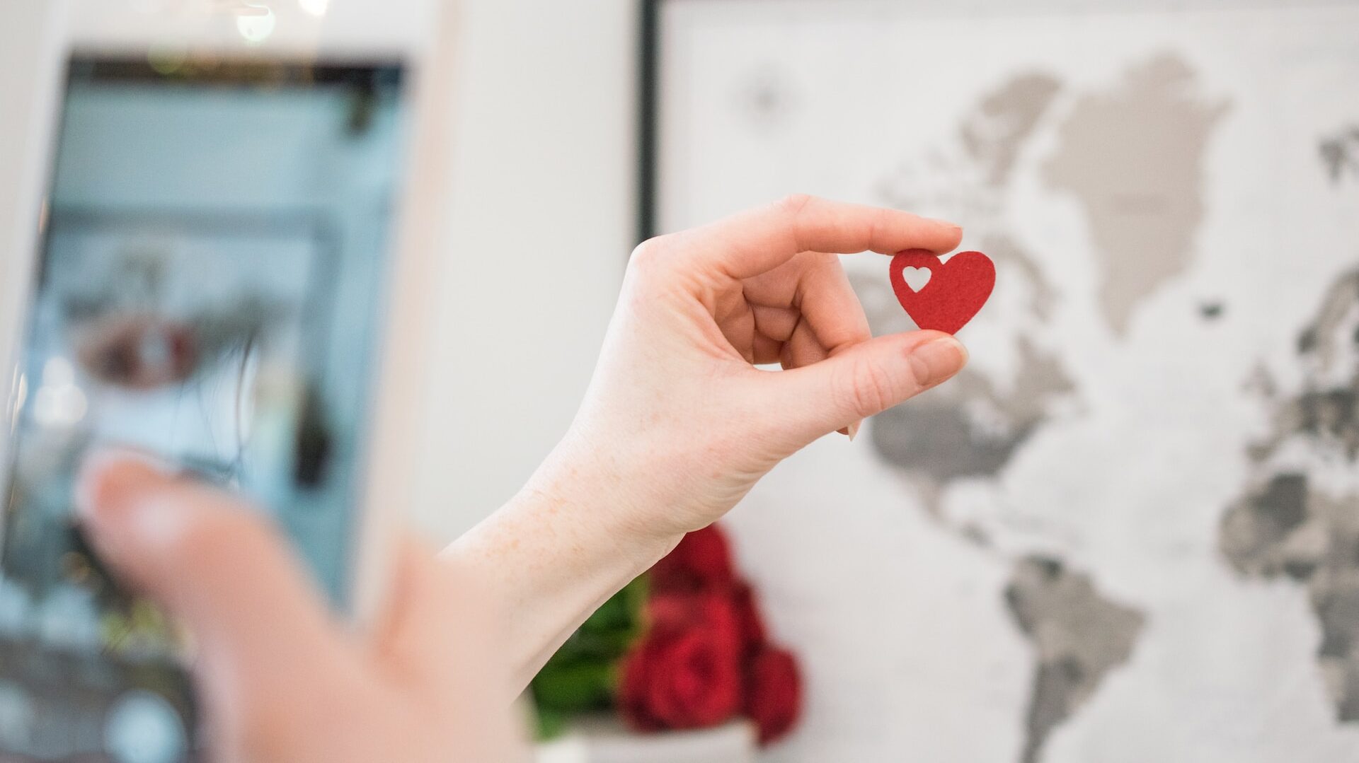 A hand holding a red paper heart in front of a map of the world, out of focus in the background.
