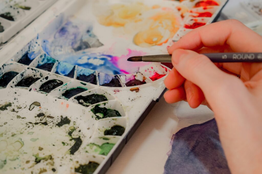 Hand holding a paintbrush above a messy watercolour tray