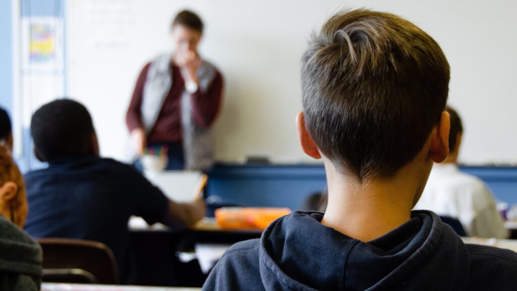a boy sat in class, viewed from the back. A teacher is visible from the blurred foreground