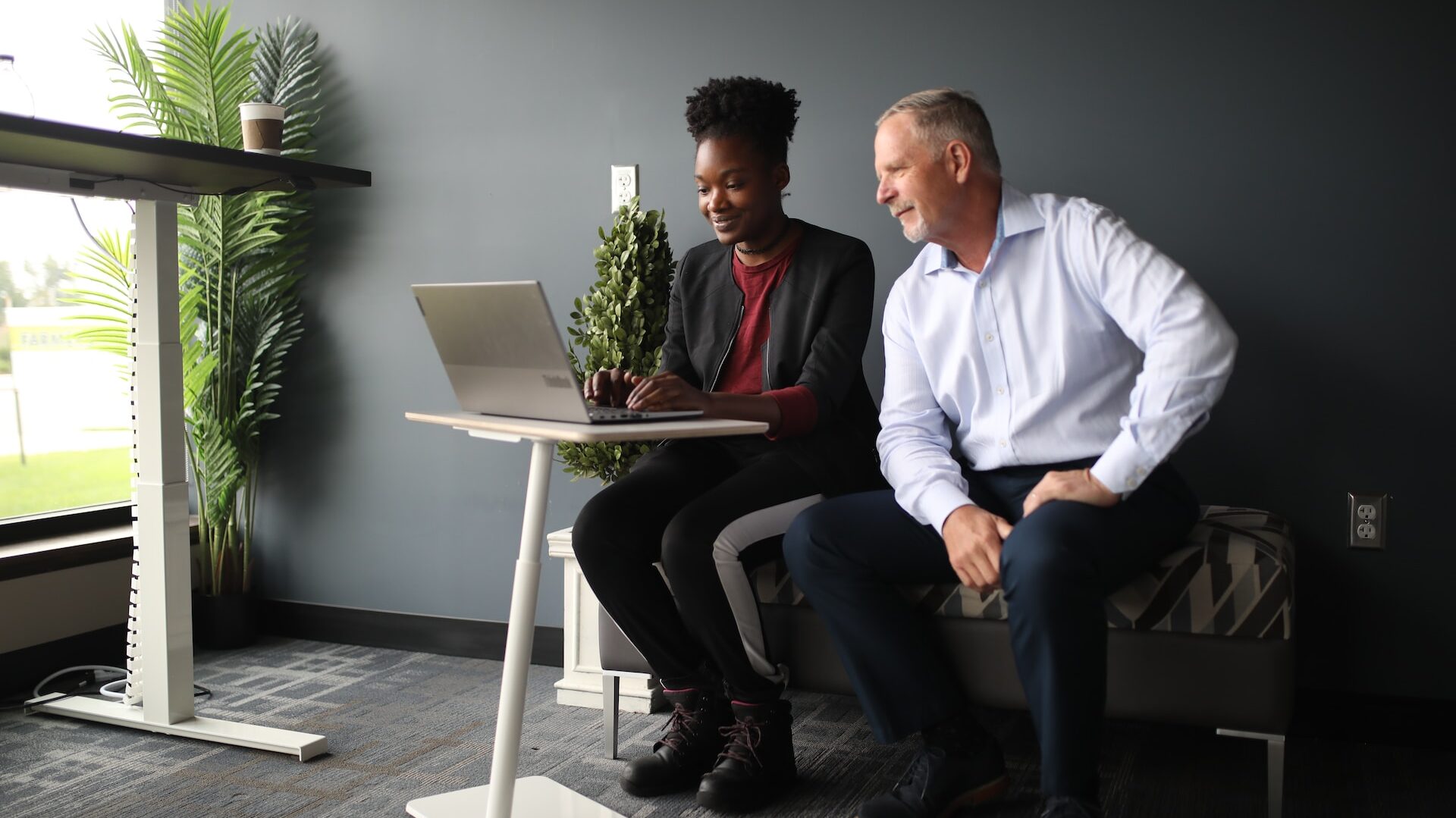 an older person sat next to a younger colleague in an open-plan office, looking at a laptop as they type 