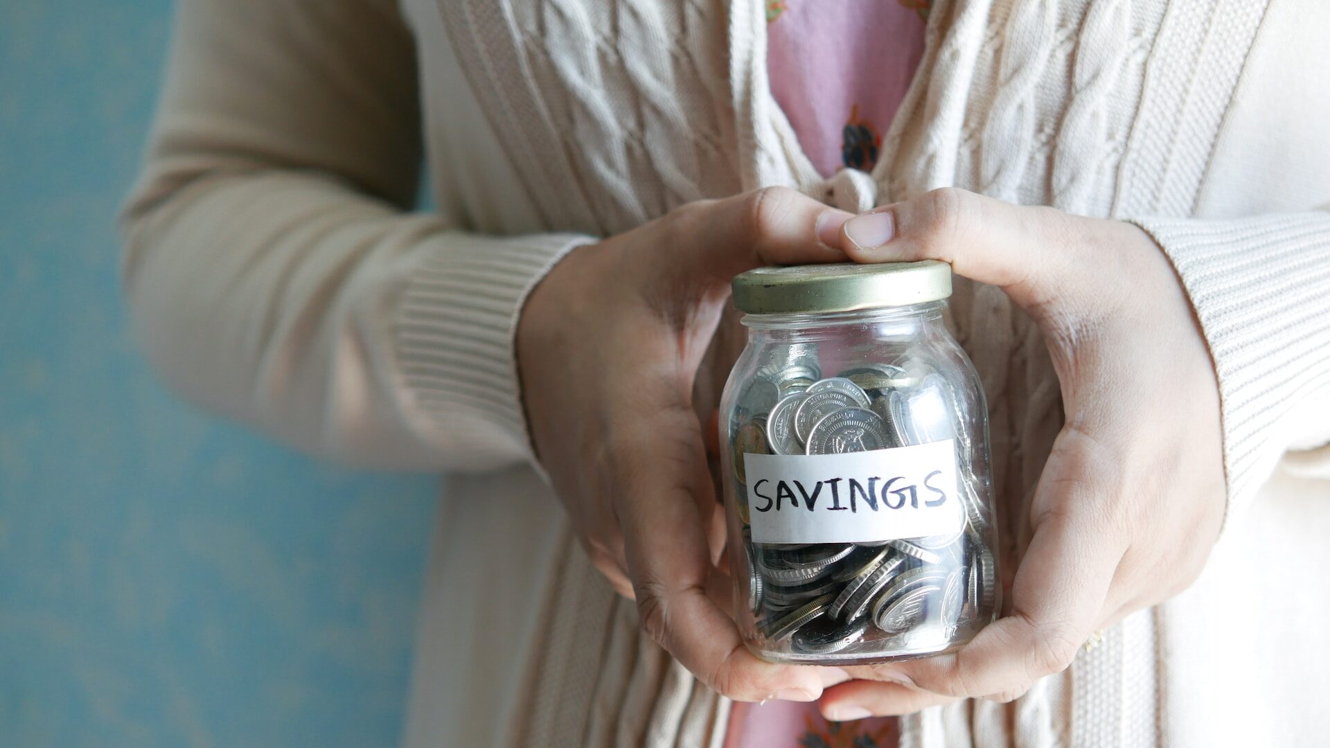 a person in a cardigan holding a jar of silver coins in their hands, labelled "savings"