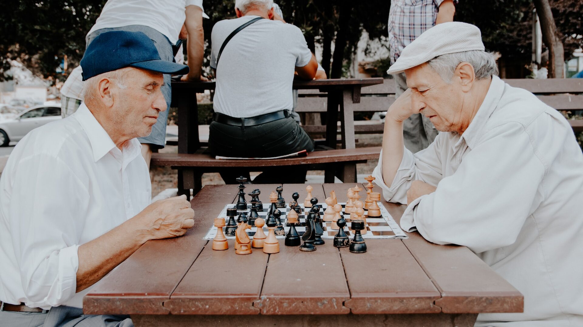 two older people in a park, both wearing caps, engaged in a game of chess